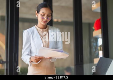 Businesswoman standing near laptop and reviewing documents in modern office, showing professional planning, financial paperwork, project management, c Stock Photo