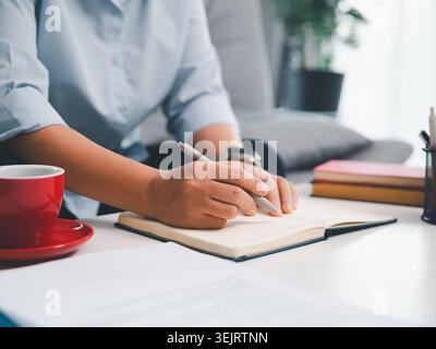 Professional writing in open notebook with red coffee cup on bright office desk, showing note taking, business planning, creative ideas, productivity, Stock Photo