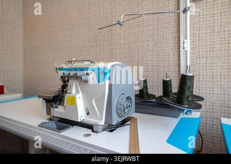 An industrial overlock sewing machine with a white and blue casing is positioned on a worktable with several spools of dark green thread on a metal st Stock Photo