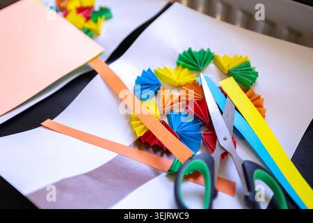 A pair of metal scissors sits on sheets of paper alongside strips of colored construction paper and small folded accordion fans as part of an art and Stock Photo