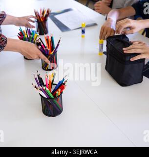 Hands of an adult teacher and elementary school children organize colored drawing pencils in black mesh holders alongside yellow glue sticks and bags Stock Photo