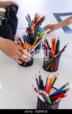 An adult and a child reach for black metal mesh cups filled with various colorful wooden drawing pencils and felt-tip pens arranged on a white table d Stock Photo