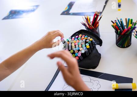 A close-up view of a child using hands to select a marker from a black fabric case filled with creative art supplies on a white table surrounded by co Stock Photo