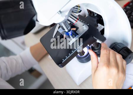 High-angle view of a laboratory technician adjusting a glass slide with a sample on an optical microscope stage under blue light illumination during a Stock Photo
