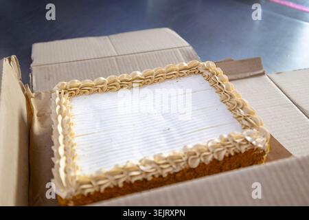 A rectangular sheet cake with white frosting and a cream-colored piped border rests inside an open cardboard box placed on a dark metal tabletop surfa Stock Photo
