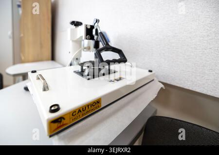 A white manual clamshell heat press machine with a prominent yellow caution hot surfaces warning sticker on the front plate rests on a grey worktable Stock Photo