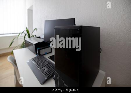 A black desktop computer tower stands on a white office desk alongside a keyboard, computer monitor, printer, and office accessories in a brightly lit Stock Photo