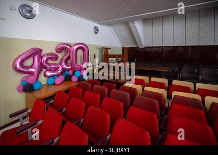 Rows of red upholstered seats fill an empty interior assembly hall where large pink foil number balloons are arranged on a wooden desk beside stacked Stock Photo