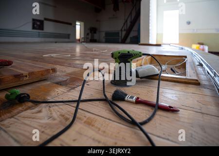 An orbital sander and a paint brush with a red handle sit on a wooden plank floor during a renovation process inside a large empty room with a stage a Stock Photo
