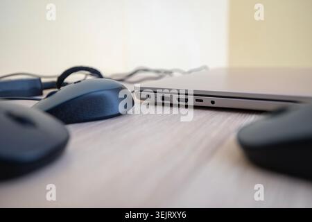 A close-up view of a modern silver laptop computer positioned on a light wooden desk surface next to multiple black computer mice with focus on the si Stock Photo