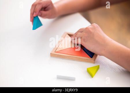 Hands of a child arrange colorful triangular wooden pieces into a square frame during a geometric puzzle activity on a white table to support cognitiv Stock Photo