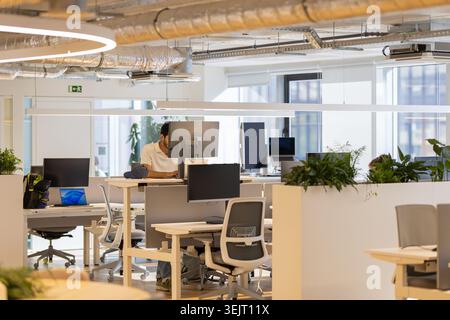 Modern office workspace featuring a male employee working at a computer desk surrounded by greenery and multiple monitors in a bright, open-plan Stock Photo