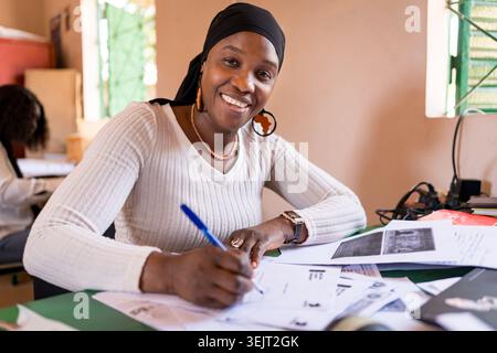 Portrait of smiling young african business woman sitting at desk in office writing and working on laptop, enterpreneurship in africa Stock Photo