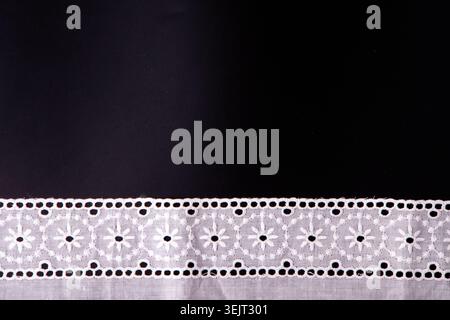 White floral lace ribbon trim on black background, delicate cotton embroidery border fabric mockup. Top view embroidered eyelet flower patterns on a black surface. Text copy space. Stock Photo