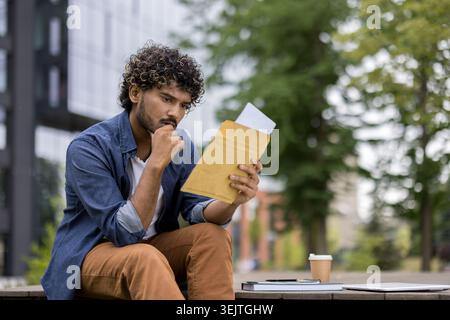 Young college student sitting outdoors on campus reading a serious letter, looking concerned and pensive about academic or financial news, uncertainty about the future Stock Photo