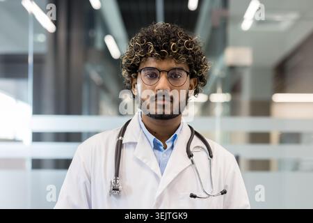 Young indian male doctor, wearing a white lab coat and stethoscope, standing professionally, looking directly at the camera in a modern hospital or clinic environment Stock Photo