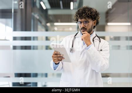 Male indian doctor wearing a white lab coat and stethoscope, standing in a clinic setting, deeply concentrating while reviewing information on a digital tablet and holding his chin Stock Photo
