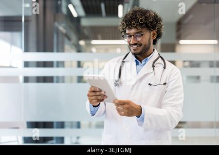 Young male doctor wearing a white lab coat and stethoscope, smiling while confidently using a digital tablet, representing modern healthcare, medical technology, and innovation in a clinic setting Stock Photo