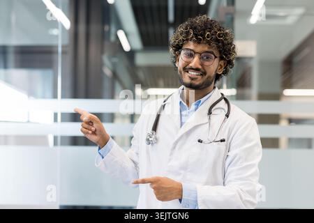 Young indian male doctor smiling and confidently pointing with both hands, presenting medical or healthcare information in a modern hospital or clinic setting Stock Photo