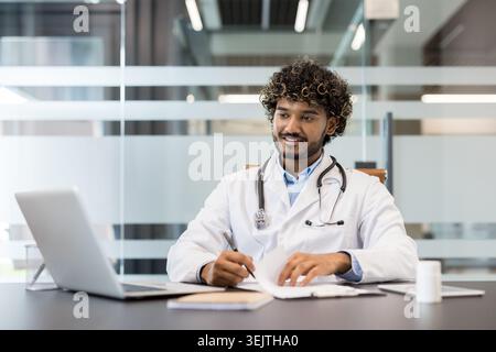 Young indian male doctor in lab coat with stethoscope, smiling and focused while writing notes and using a laptop at a modern clinic desk for telehealth consultation Stock Photo