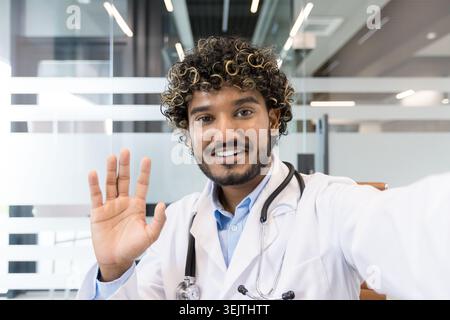 Indian male doctor with curly hair and a stethoscope, smiling and waving his hand during a virtual video call, providing remote telemedicine healthcare assistance in a modern office setting Stock Photo