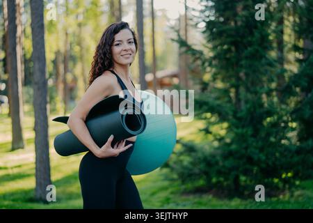 Sideways shot of beautiful curly brunette woman carries rolled up karemat and fitness ball, dressed in active wear, poses in forest or park, has fitness training outdoor, leads active lifestyle Stock Photo