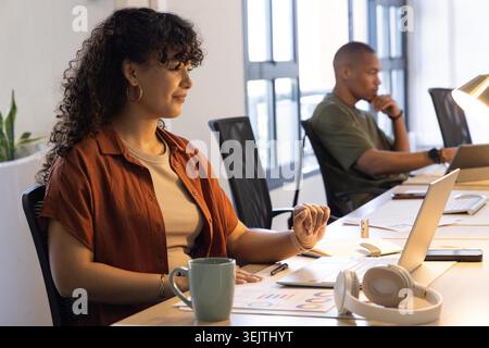 Diverse coworkers working at long wooden desk in open-plan office with laptops and charts. Colleagues, workspace, minimalist, modern, daylight, collaboration, productivity Stock Photo
