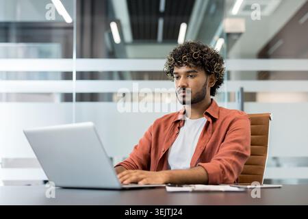 Young indian man with curly hair intently typing on a laptop, concentrating on his work at a desk in a contemporary co-working office space with glass partitions Stock Photo