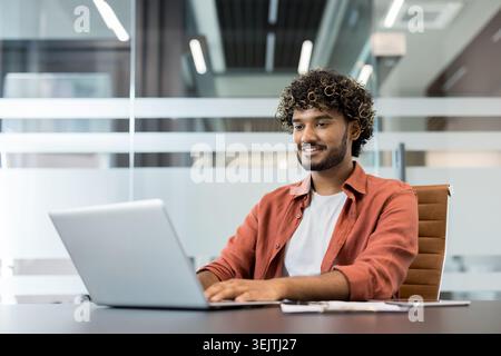 Young indian man with curly hair smiling and focused, working remotely on his laptop in a contemporary glass office, embracing productivity and professional tasks Stock Photo
