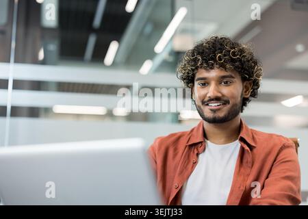 Indian man in a casual shirt smiling and looking at the camera while using a laptop, representing a positive and engaged professional in a modern office environment Stock Photo