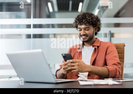 Smiling young indian man working in a modern office, using a smartphone while a laptop is on the desk, illustrating connectivity, communication, and digital technology Stock Photo