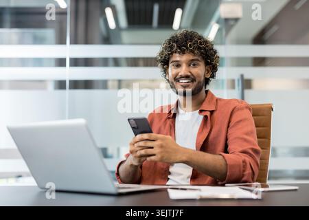 Young indian man with curly hair and beard smiling for camera, sitting at a modern office desk using smartphone while working with laptop and digital tablet Stock Photo