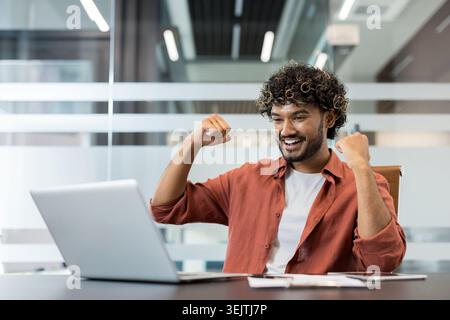 Young indian man with curly hair celebrating success and achievement, looking happy and excited while looking at laptop screen in a modern office setup Stock Photo