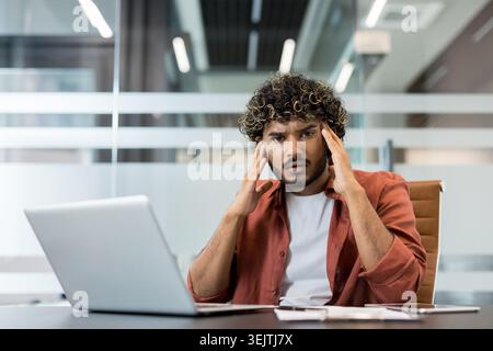 Young man experiencing headache or migraine, feeling overworked, stressed, and frustrated while sitting at a desk with a laptop in a modern office, indicating work pressure and mental health issues Stock Photo