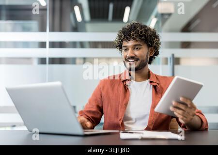 Young indian man with curly hair smiling and looking away, using a laptop and a tablet simultaneously, embodying modern multitasking and digital work in a contemporary office setting Stock Photo