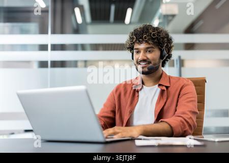 Young man wearing a headset and smiling while interacting with a client on a laptop, working as a call center agent or providing online customer service Stock Photo