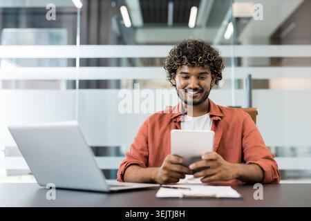 Young man with curly hair smiling while working on a digital tablet at a desk, with a laptop and documents, in a contemporary glass office environment Stock Photo