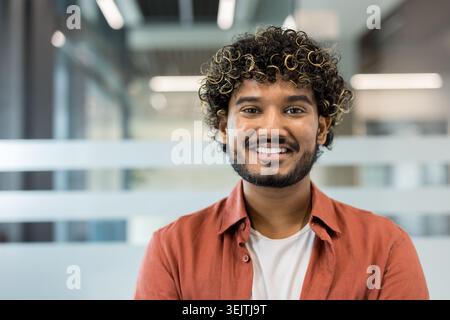 Young indian man with curly hair and beard smiling confidently in a modern office headshot, conveying friendly professionalism and upbeat startup or corporate energy Stock Photo