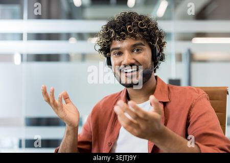Young male technical support agent with headset smiling at camera in modern office, providing customer service and remote assistance during a video call or phone consultation Stock Photo