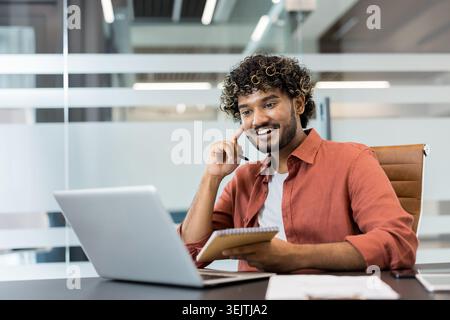 Young indian man with curly hair smiling confidently while working on a laptop and jotting notes, productive during a virtual meeting in a modern office setting Stock Photo