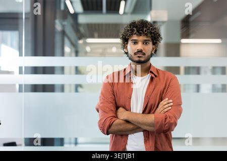 Young indian man with curly hair and beard stands arms crossed in a modern office, projecting confidence and leadership in smart-casual attire, focused and professional Stock Photo