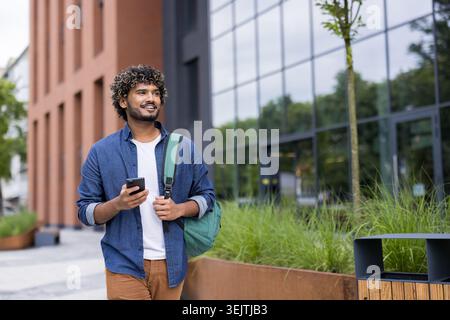Young man with a backpack walking confidently on a university campus, smiling while holding a smartphone and looking forward, embodying student life and educational aspirations Stock Photo