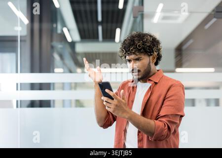 Young indian man with curly hair looking at smartphone with confused and frustrated facial expression, gesturing with hand, having problem or mistake in modern office Stock Photo