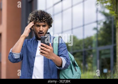 Young indian student reacting with shock and surprise to bad news on a mobile phone, expressing disbelief while standing outdoors on a university campus Stock Photo