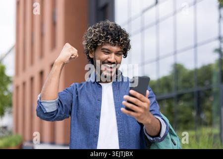 Happy young man with curly hair celebrating success and joyfully pumping his fist while looking at a smartphone, standing outdoors in an urban environment with a modern building in the background Stock Photo