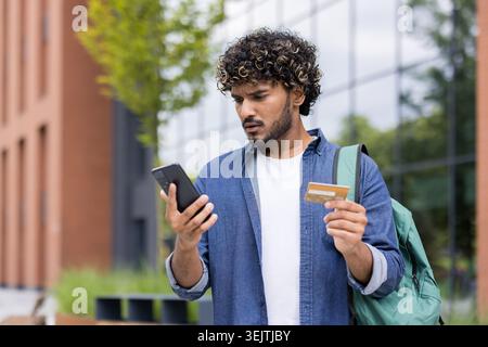 Young man standing outdoors, frustrated and confused while trying to complete a mobile banking or online payment with smartphone and credit card, facing technical or data entry errors Stock Photo