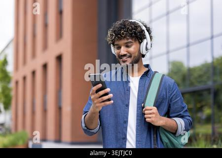 Young indian man with headphones and backpack walking on campus, smiling while checking his phone and enjoying music or a podcast, relaxed and connected in a modern university setting Stock Photo