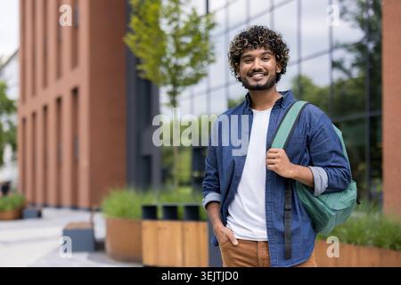 Young indian male student with curly hair smiling and looking at camera, carrying a backpack while standing outside a university building, representing education and lifestyle Stock Photo