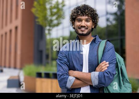 Young indian man with curly hair and a backpack smiling confidently with arms crossed, standing outdoors on a modern university campus, representing education and successful student life Stock Photo