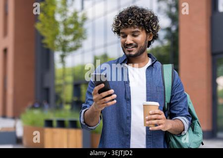 Young indian student standing on urban street, smiling while using smartphone, holding coffee and backpack, representing modern mobile learning, social networking and city lifestyle Stock Photo